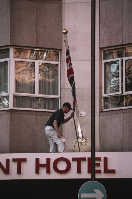 A man wearing dark clothing and light-colored pants is cleaning or repairing a tall flagpole outside a building, which appears to be a hotel. The building's exterior consists of concrete panels with l