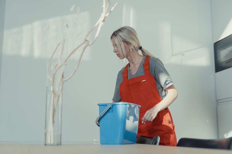 Two professional cleaners in red overalls are performing surface cleaning in a modern kitchen. The female cleaner in the foreground is vacuuming the tiled floor with a handheld vacuum cleaner, demonst